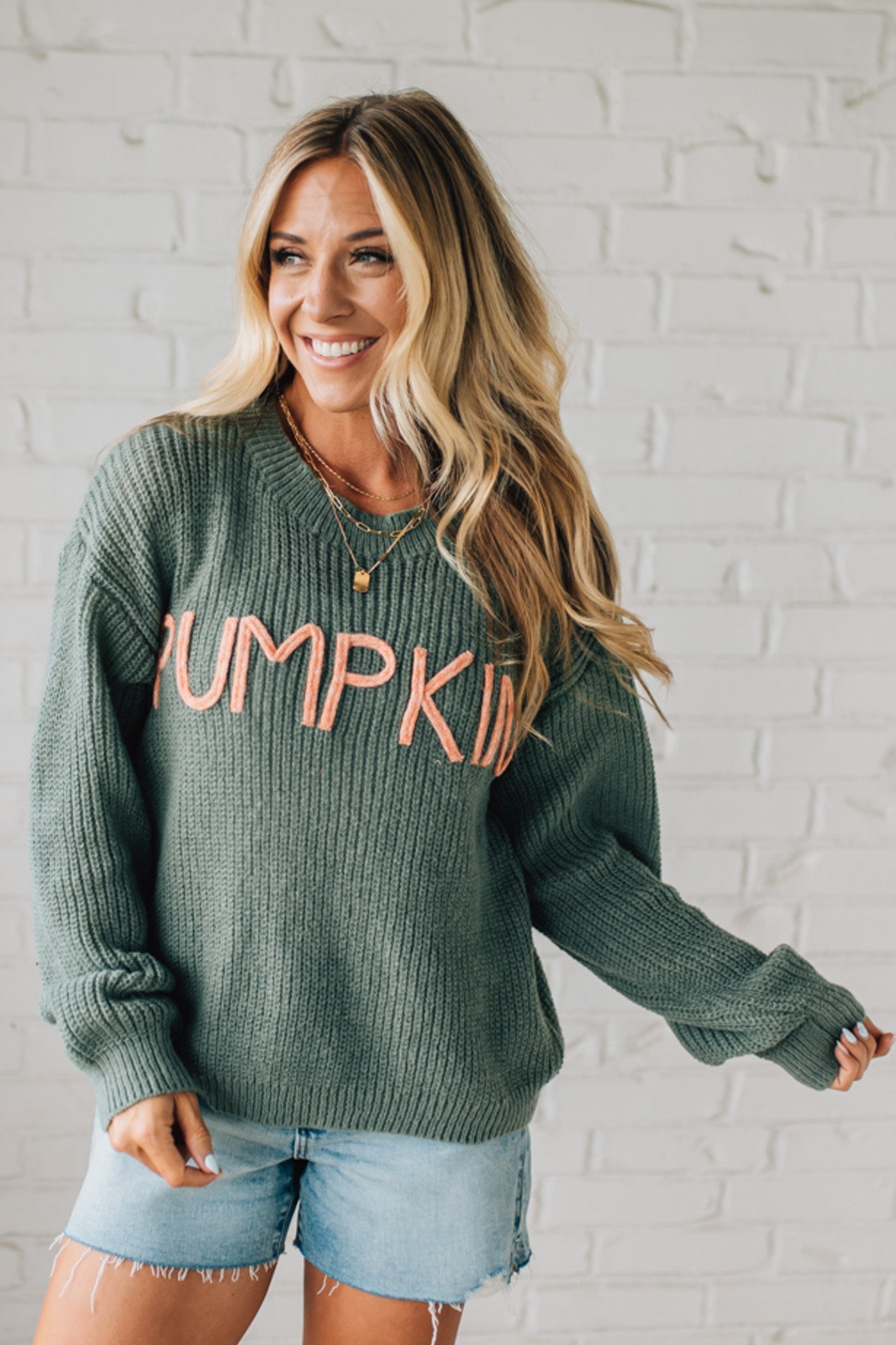 Woman wearing a green sweater with 'Pumpkin' text against a white brick wall.
