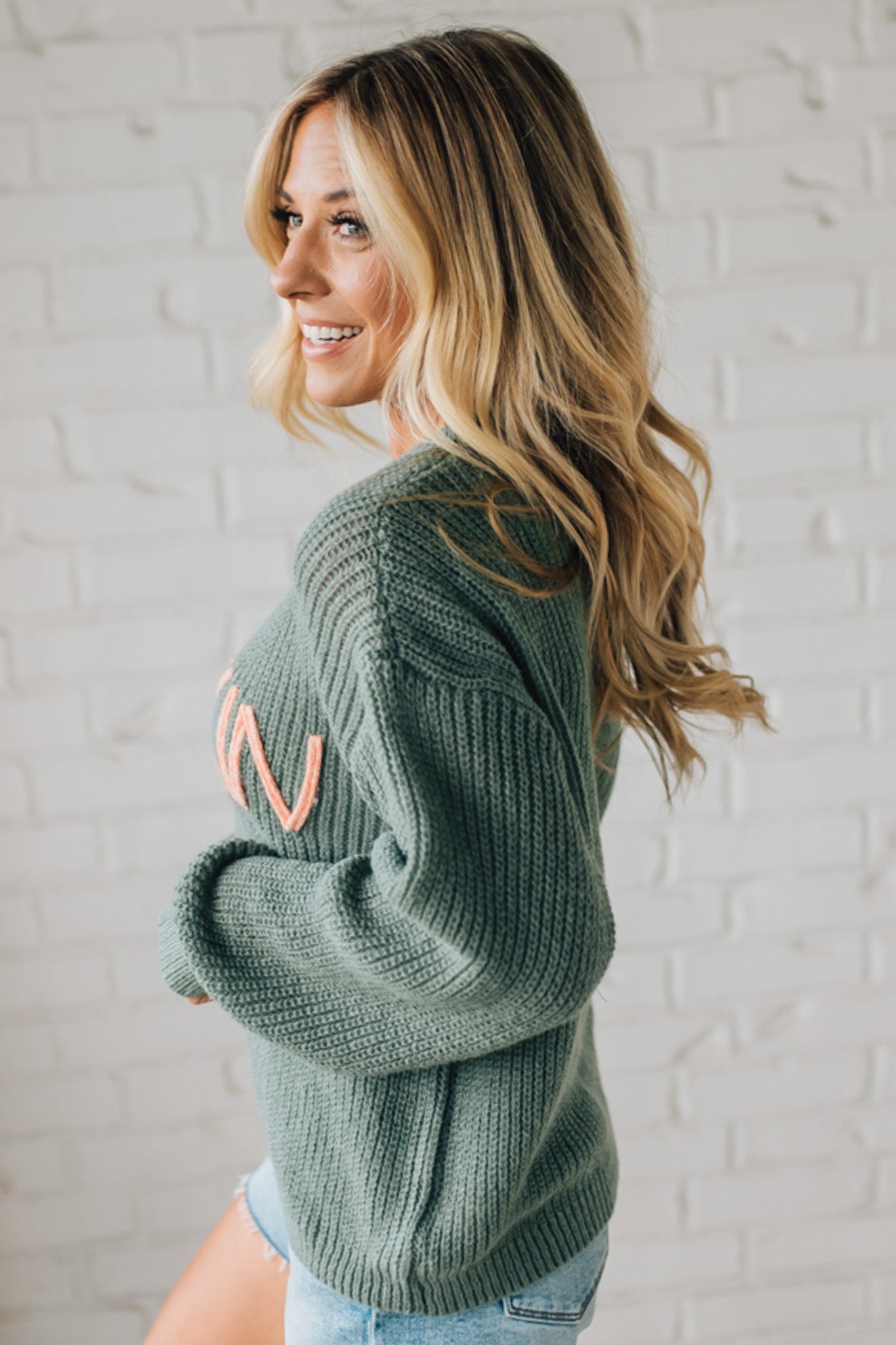 Woman wearing a green sweater with a pink letter 'N' on a white brick wall background