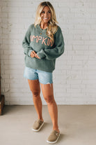 Woman wearing a green sweater with 'Pumpkin' text, light blue shorts, and beige shoes against a white brick wall.