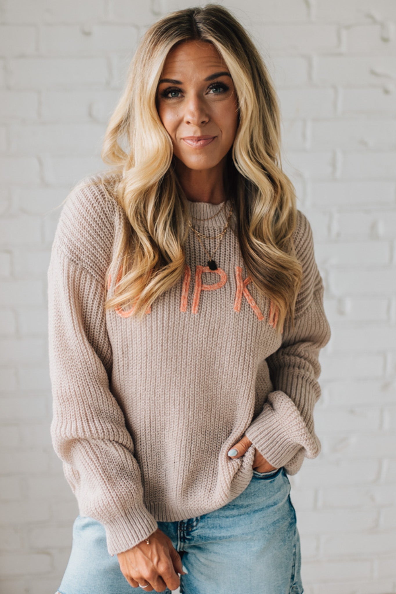 Woman wearing a beige sweater with 'IPK' text against a white brick wall