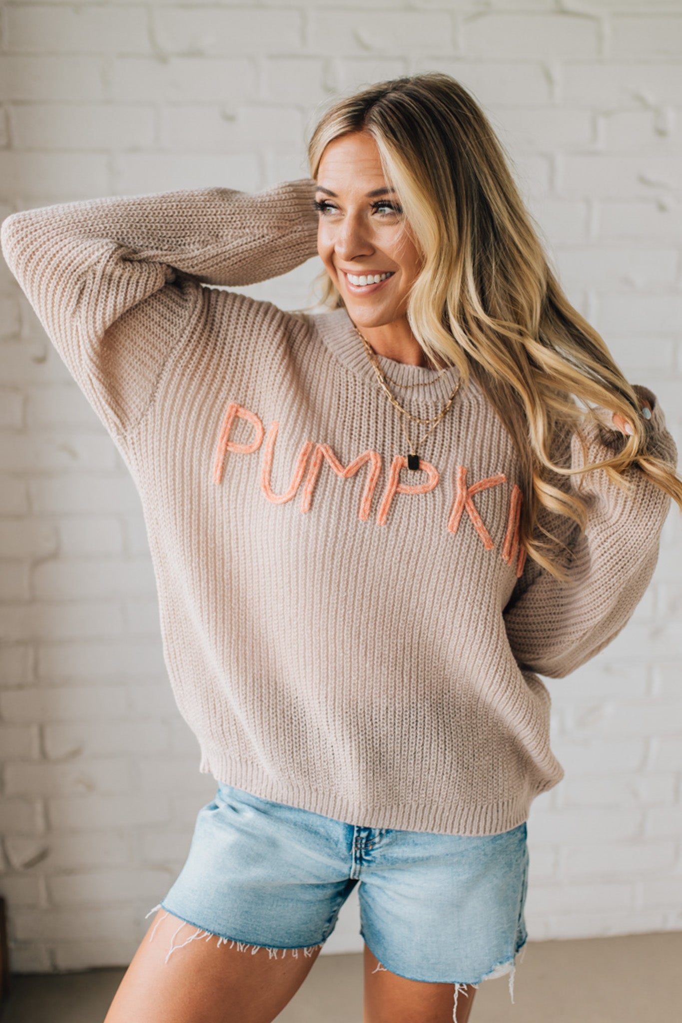 Woman wearing a beige sweater with 'PUMPKIN' in pink text, standing against a white brick wall.