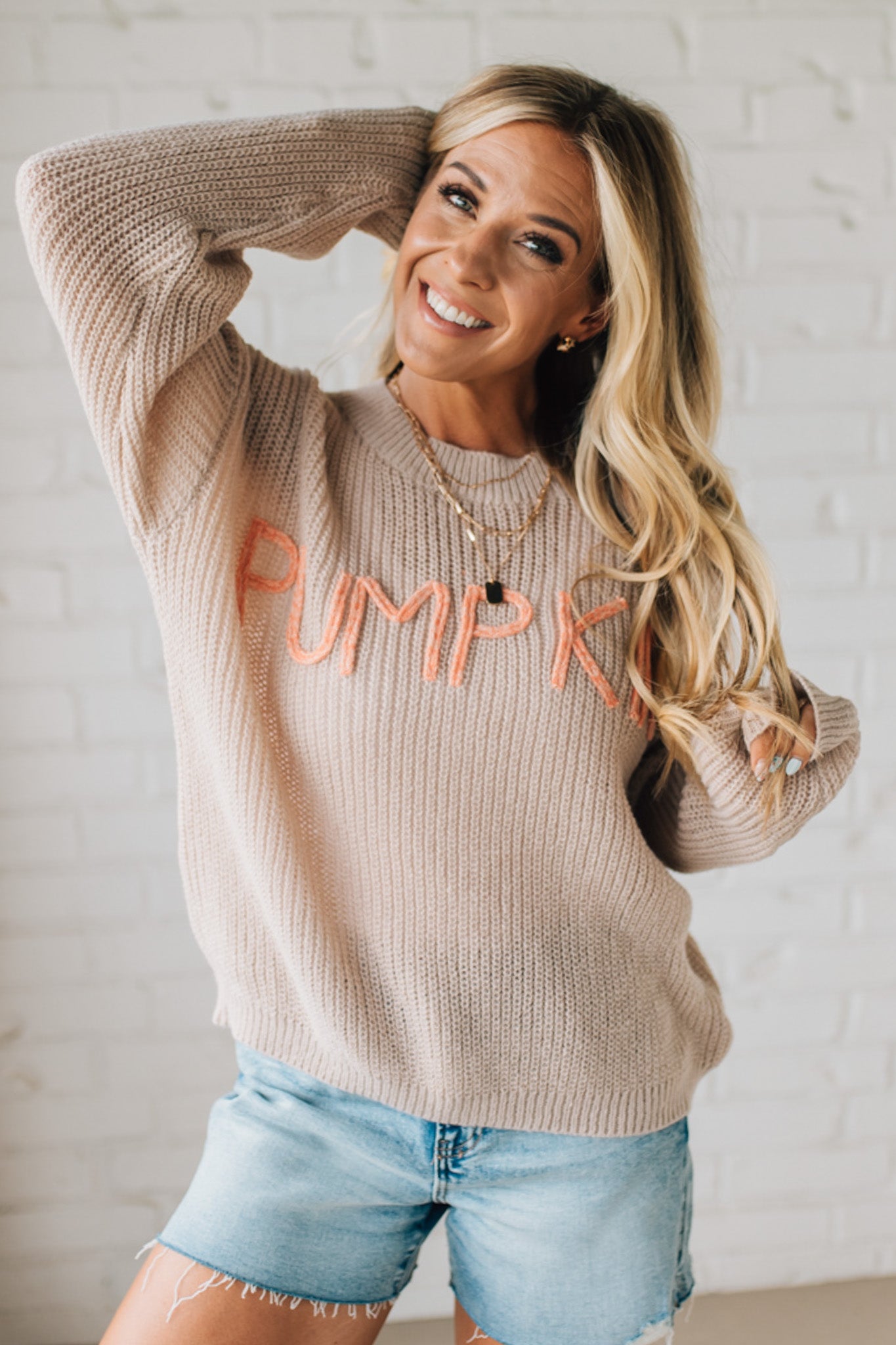 Woman wearing a beige sweater with 'Pumpkin' text, standing against a white brick wall.