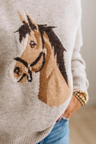 BLONDE WOMAN IN DARK JEANS WEARING A HORSE JACQUARD, STITCH TRIM ACCENT, LONG SLEEVE, CREW NECK PULLOVER SWEATER WITH A HORSE ON THE FRONT, AND WEARING A BIG BELT BUCKLE TOO.
