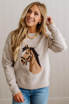 BLONDE WOMAN IN DARK JEANS WEARING A HORSE JACQUARD, STITCH TRIM ACCENT, LONG SLEEVE, CREW NECK PULLOVER SWEATER WITH A HORSE ON THE FRONT, AND WEARING A BIG BELT BUCKLE TOO.
