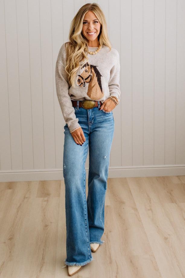 BLONDE WOMAN IN DARK JEANS WEARING A HORSE JACQUARD, STITCH TRIM ACCENT, LONG SLEEVE, CREW NECK PULLOVER SWEATER WITH A HORSE ON THE FRONT, AND WEARING A BIG BELT BUCKLE TOO.
