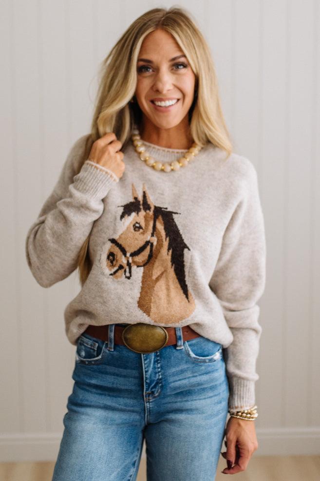 BLONDE WOMAN IN DARK JEANS WEARING A HORSE JACQUARD, STITCH TRIM ACCENT, LONG SLEEVE, CREW NECK PULLOVER SWEATER WITH A HORSE ON THE FRONT, AND WEARING A BIG BELT BUCKLE TOO.
