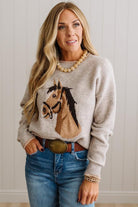 BLONDE WOMAN IN DARK JEANS WEARING A HORSE JACQUARD, STITCH TRIM ACCENT, LONG SLEEVE, CREW NECK PULLOVER SWEATER WITH A HORSE ON THE FRONT, AND WEARING A BIG BELT BUCKLE TOO.
