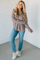 Woman wearing a brown sweater, blue jeans, and white sneakers against a white brick wall.