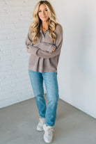 Woman wearing a brown sweater, blue jeans, and white sneakers against a white brick wall.