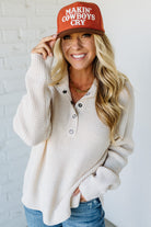 Woman wearing a beige cardigan and jeans, holding a red cap with text against a white brick wall.