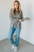 Woman wearing a gray sweater and blue jeans against a white brick wall.
