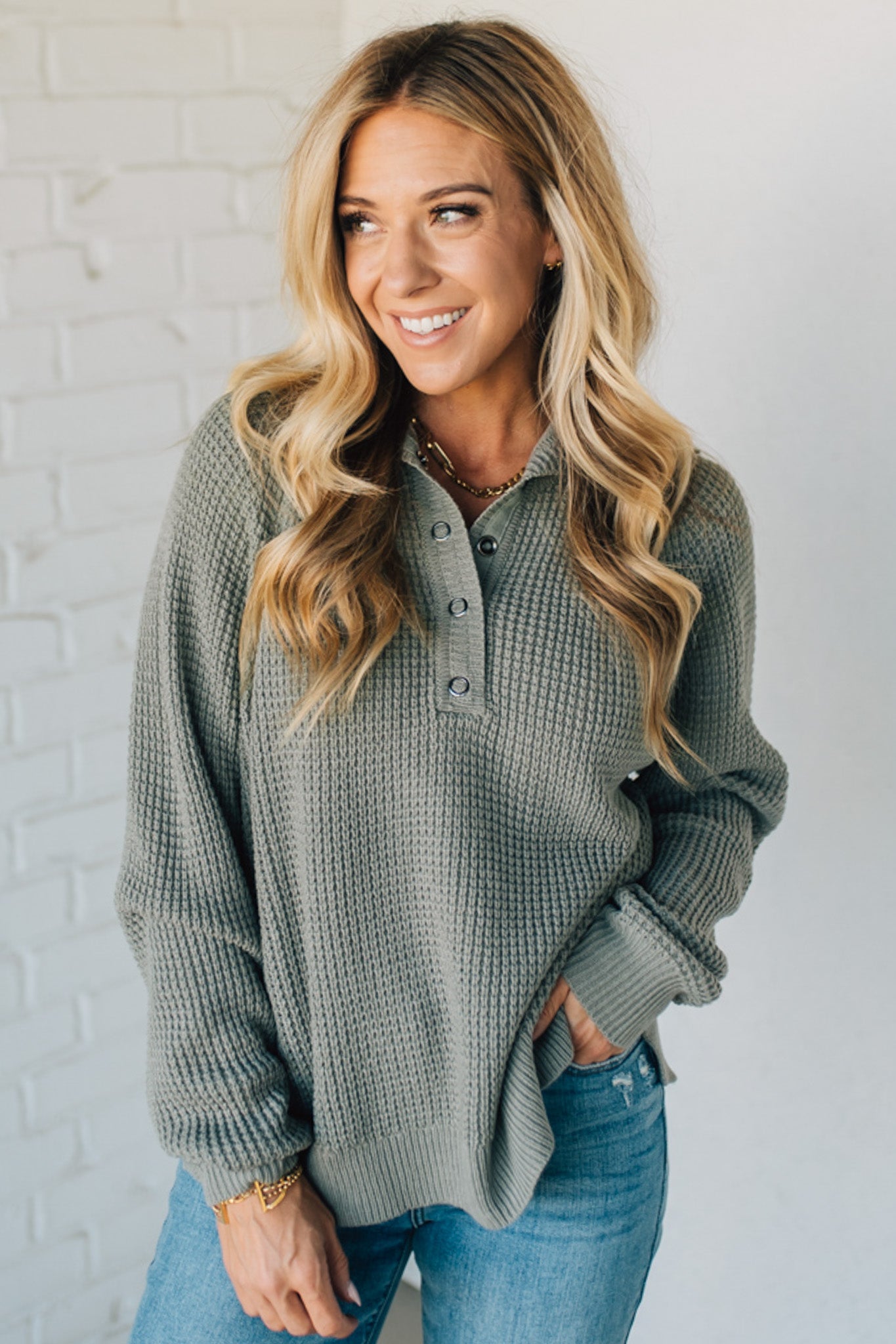 Woman wearing a green waffle knit sweater and blue jeans against a white brick wall.