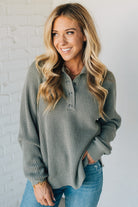 Woman wearing a green waffle knit sweater and blue jeans against a white brick wall.