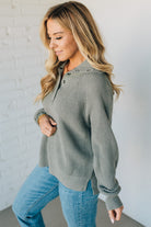 Woman wearing a green sweater and blue jeans against a white brick wall.