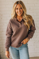 Woman wearing a brown quilted sweater and blue jeans against a white brick wall.