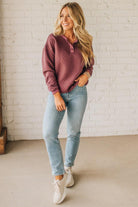Woman wearing a mauve sweater and light blue jeans standing against a white brick wall.