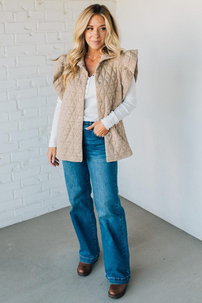 Woman wearing a beige quilted vest, white shirt, blue jeans, and brown shoes against a white brick wall.