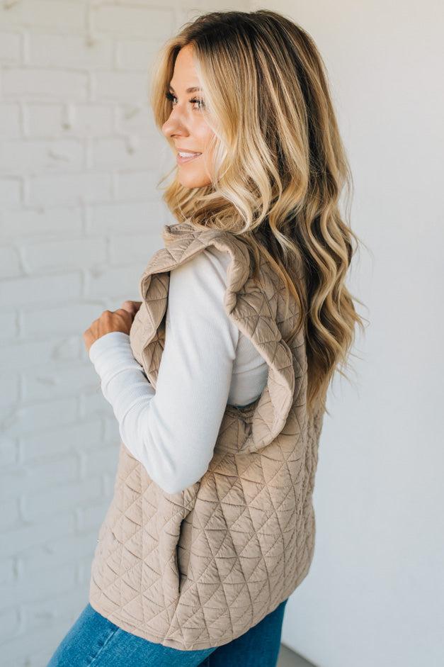 Woman wearing a beige quilted vest against a white brick wall