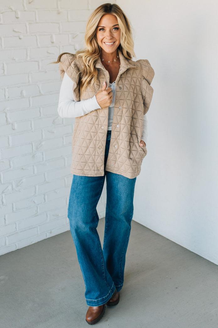 Woman wearing a beige quilted vest, white shirt, blue jeans, and brown shoes against a white brick wall.