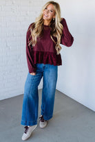 Woman wearing a burgundy ruffled top and blue jeans against a white brick wall.
