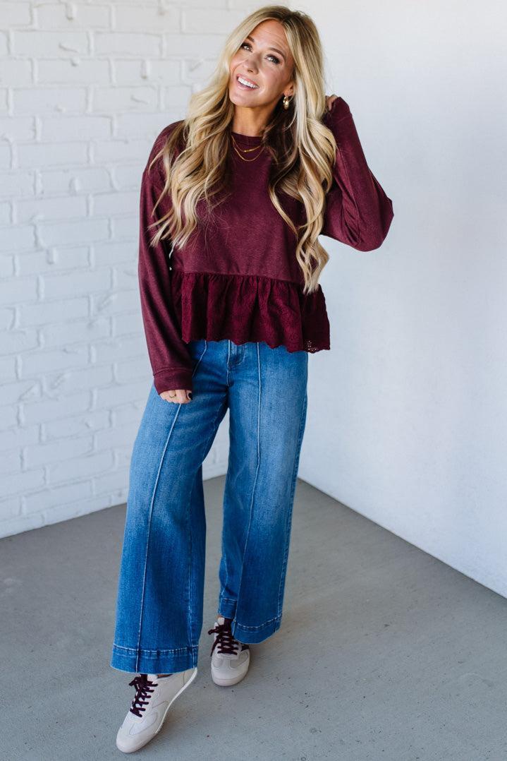 Woman wearing a burgundy ruffled top and blue jeans against a white brick wall.