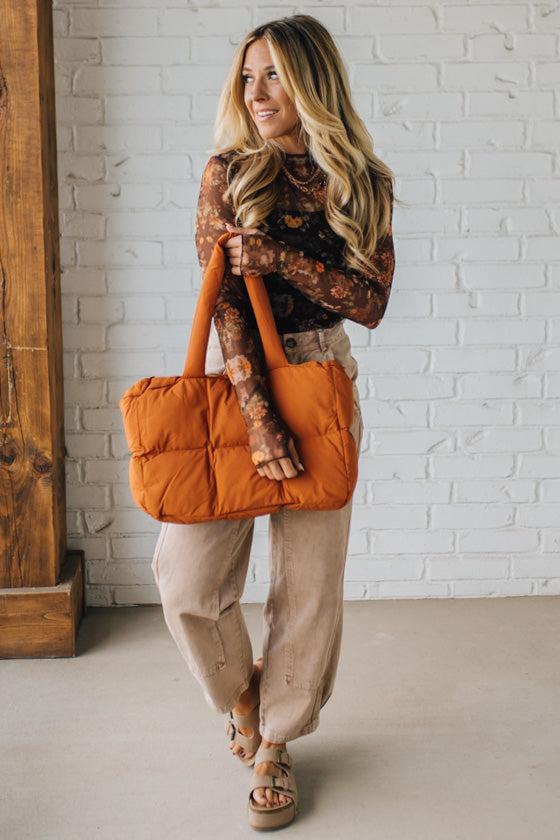 Woman holding an orange puffer bag against a white brick wall.