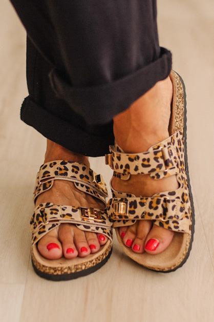 Close up of women's leoapard print double strap sandals with gold buckles on feet with painted orange toenails, and black cuffed pants. 