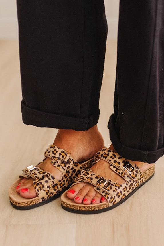 Close up of women's leopard print double strap sandals with gold buckles on feet with painted orange toenails, and black cuffed pants. 