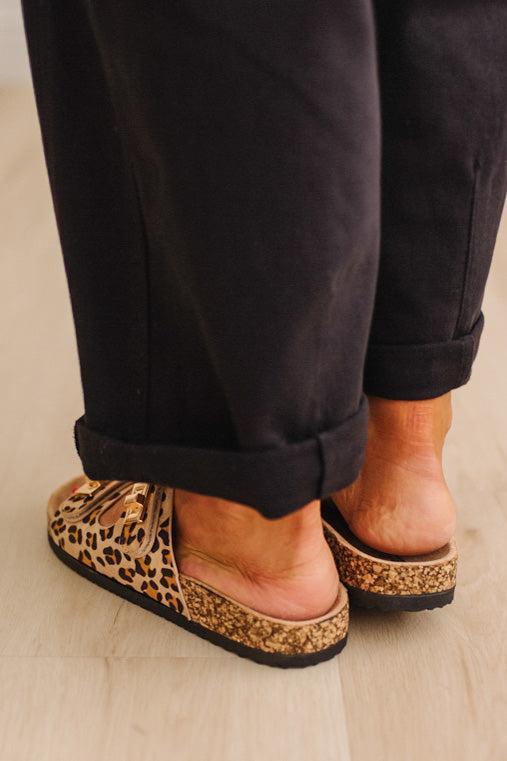 Close up of women's leopard print double strap sandals with gold buckles on feet with painted orange toenails, and black cuffed pants. 