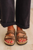 A close-up of a person wearing brown double-strap cork sandals with gold-tone buckles and black trim, paired with dark cuffed pants on a light wood floor.