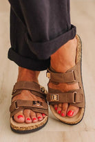 A close-up of a person wearing brown double-strap sandals with gold-tone hardware and cork-look soles, paired with dark cuffed pants on a light wood floor.