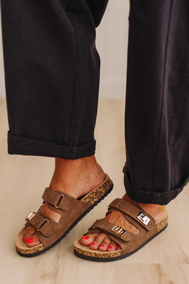 A person wearing brown double-strap slide sandals with cork-look soles and black pants, standing on a light wood floor.