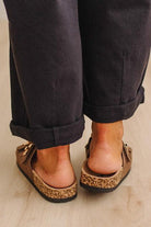 A close-up rear view of a person wearing brown leather clogs with cork-look soles and gold-tone hardware, paired with dark cuffed pants on a light wood floor.