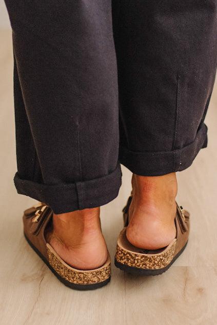 A close-up rear view of a person wearing brown leather clogs with cork-look soles and gold-tone hardware, paired with dark cuffed pants on a light wood floor.
