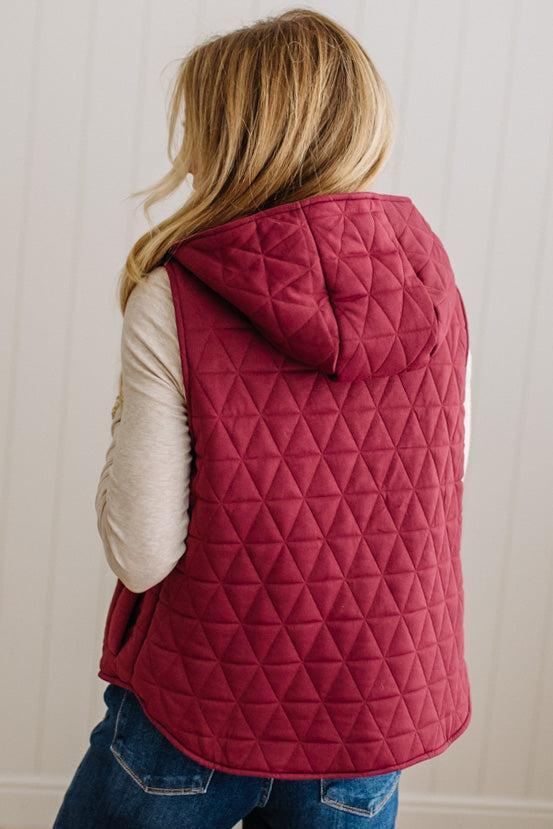 A woman shown from the back wearing a quilted hooded vest in a deep berry pink color, layered over a cream long-sleeve shirt and blue jeans, against a white paneled wall.