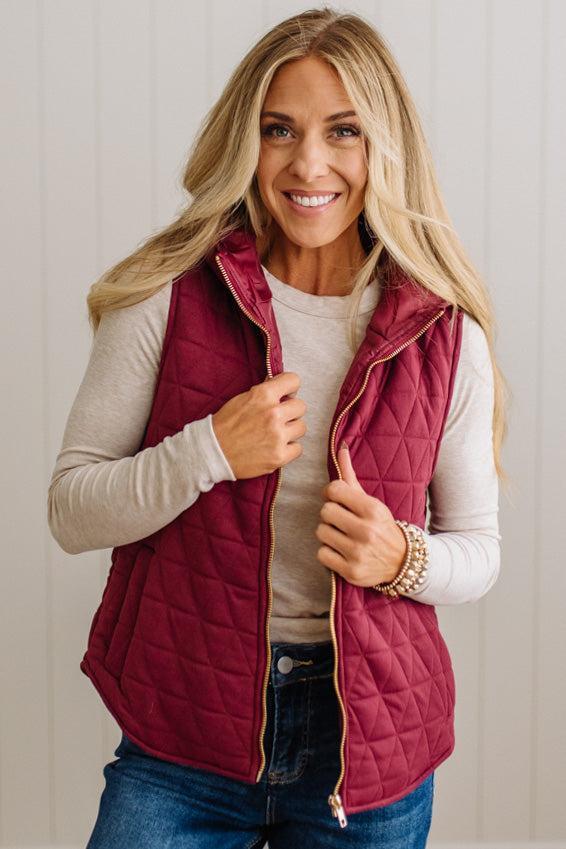 A smiling blonde woman wearing a quilted burgundy puffer vest over a light beige long-sleeve shirt and blue jeans, standing against a white paneled wall.