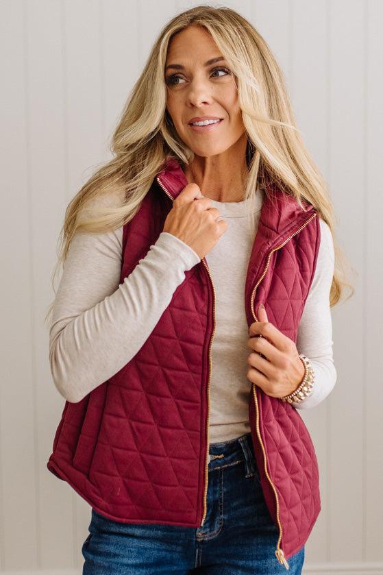 A woman wearing a quilted sleeveless vest in deep burgundy with a gold zipper, layered over a cream long-sleeve shirt and blue jeans, standing against a white paneled wall.