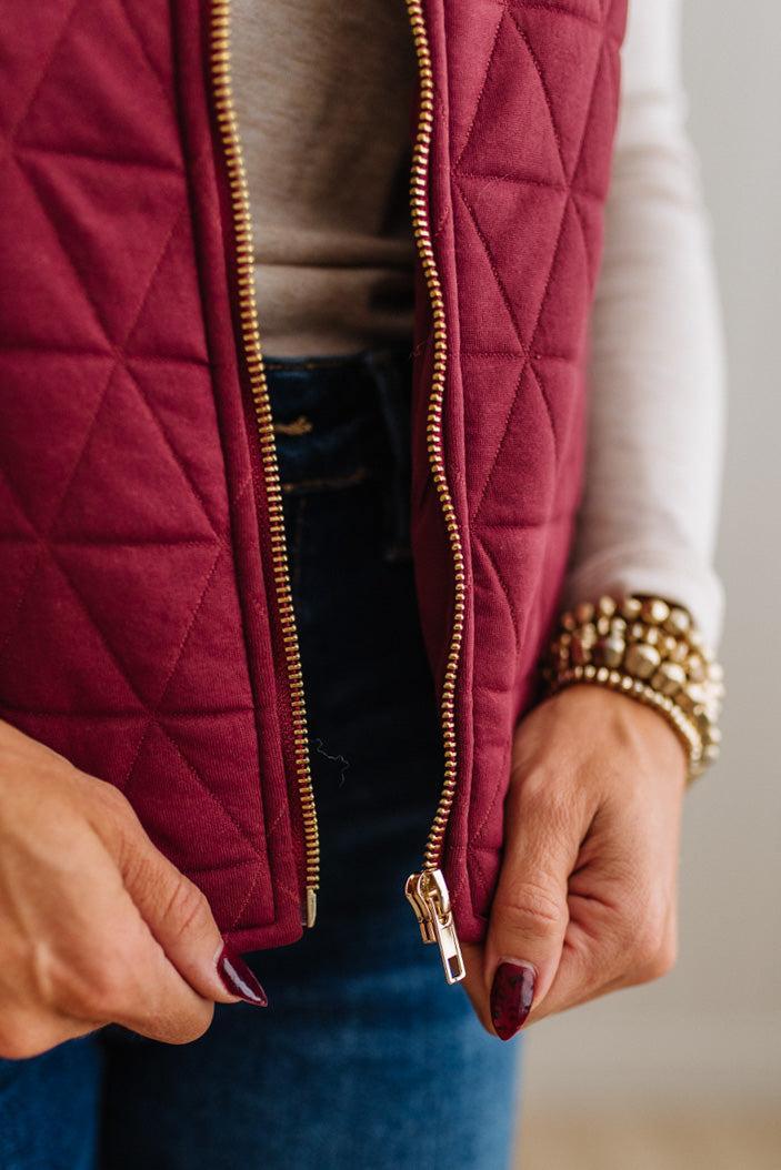 Close-up of a person wearing a quilted burgundy vest over a beige top and blue jeans, with both hands holding the zipper open. The person is also wearing stacked gold bracelets.