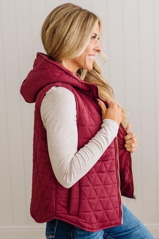 A woman wearing a quilted sleeveless hooded vest in deep burgundy over a cream long-sleeve shirt and blue jeans, shown from the side against a light paneled wall.