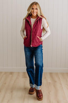 A woman modeling a sleeveless quilted burgundy vest over a cream long-sleeve top, paired with blue jeans and brown slip-on shoes, standing against a white paneled wall and light wood floor.