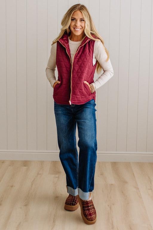 A woman modeling a sleeveless quilted burgundy vest over a cream long-sleeve top, paired with blue jeans and brown slip-on shoes, standing against a white paneled wall and light wood floor.
