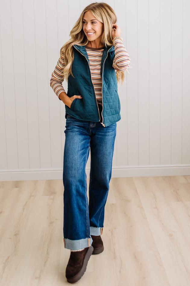 A woman modeling a teal quilted zip-front vest over a beige and brown striped long-sleeve shirt, paired with blue jeans and brown boots, standing in a room with white paneled walls and light wood floors.