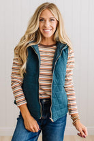 A smiling blonde woman models a teal quilted zip-front vest over a striped long-sleeve shirt and blue jeans, standing against a light paneled wall.