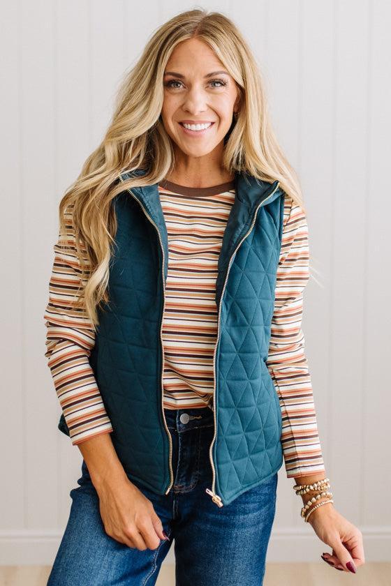 A smiling blonde woman models a teal quilted zip-front vest over a striped long-sleeve shirt and blue jeans, standing against a light paneled wall.