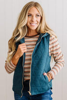 A smiling blonde woman wearing a teal quilted zip-front vest over a striped long-sleeve shirt and blue jeans, standing against a light paneled wall.