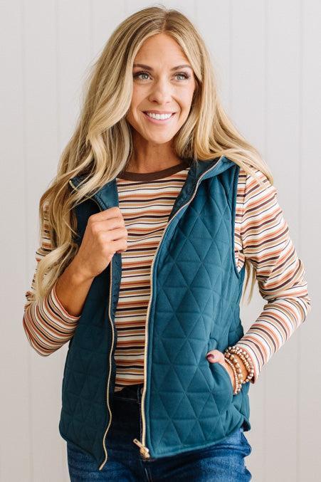 A smiling blonde woman wearing a teal quilted zip-front vest over a striped long-sleeve shirt and blue jeans, standing against a light paneled wall.