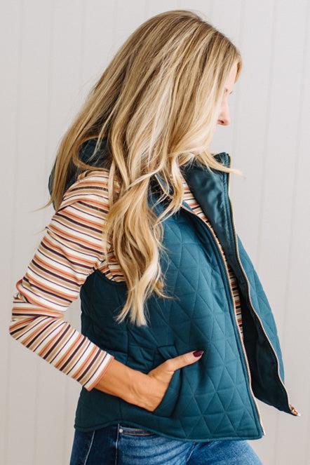 A woman shown from the side and back wearing a dark teal quilted puffer vest over a striped long-sleeve shirt and blue jeans, standing against a white paneled wall.