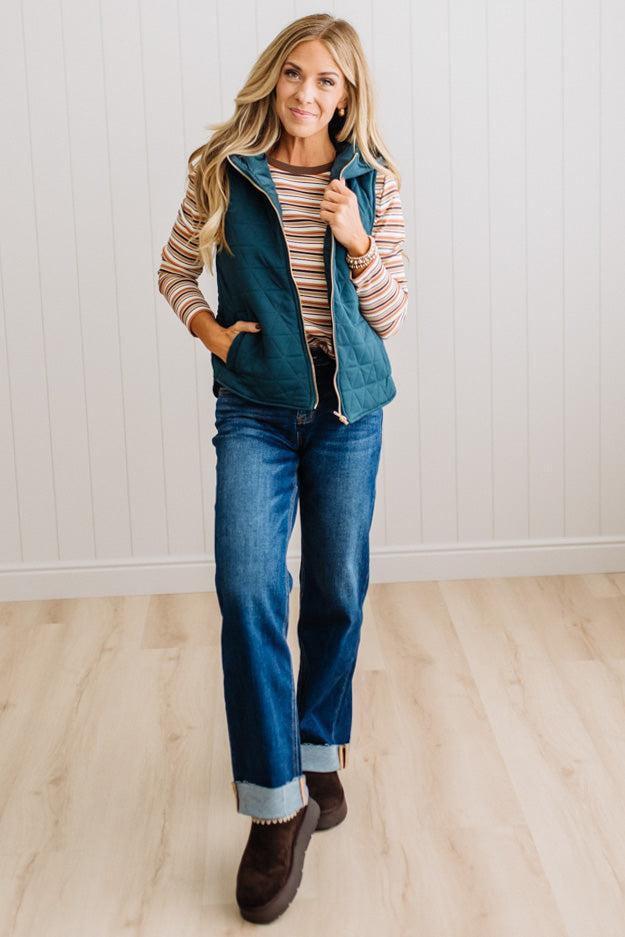 A woman modeling a teal quilted zip-up vest over a striped long-sleeve shirt, paired with blue jeans and brown boots, standing in a room with light wood floors and white paneled walls.