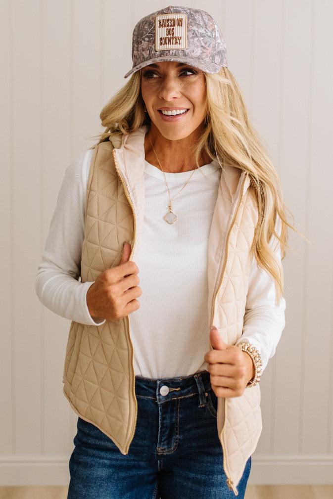 A woman wearing a beige quilted vest over a white long-sleeve top, blue jeans, and a camouflage baseball cap with a patch that reads 'BORN IN THE COUNTRY', standing against a light paneled wall.