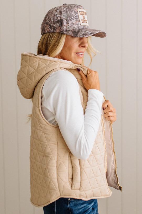 A woman wearing a beige quilted hooded vest over a white long-sleeve top and blue jeans, with a camouflage baseball cap. She is shown from the side against a light beige paneled wall.